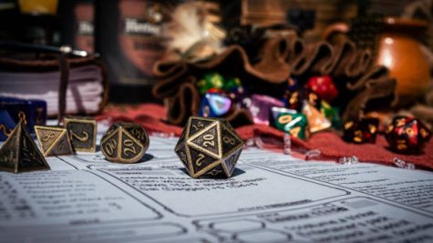 Image of a brass 20-sided die on a character sheet in the sun. In the background are a dice bag and a notebook